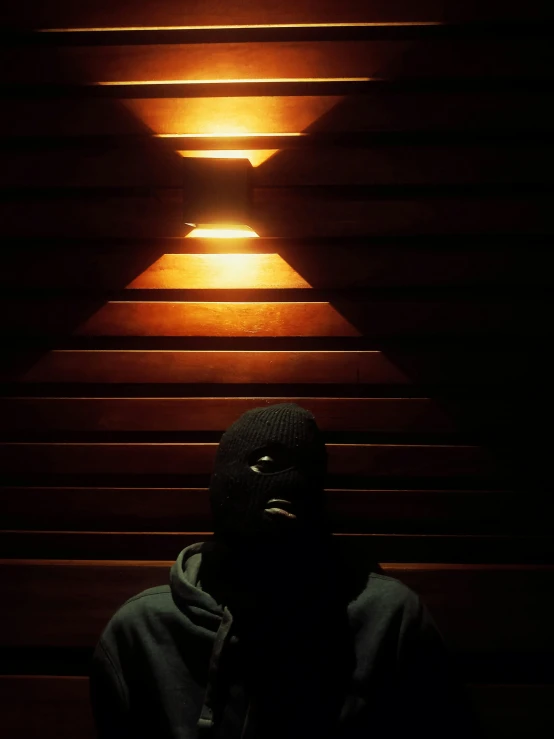 a man in a hoodie standing in front of stairs, by Attila Meszlenyi, dark skinned, cabin lights, dark visor covering face, trending photo