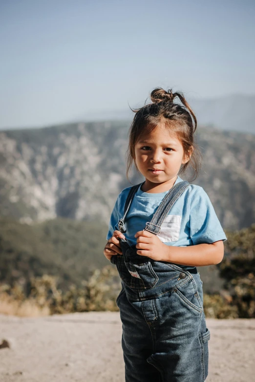 a little girl standing on top of a dirt road, on top of a mountain, wearing blue jean overalls, alanis guillen, looking serious