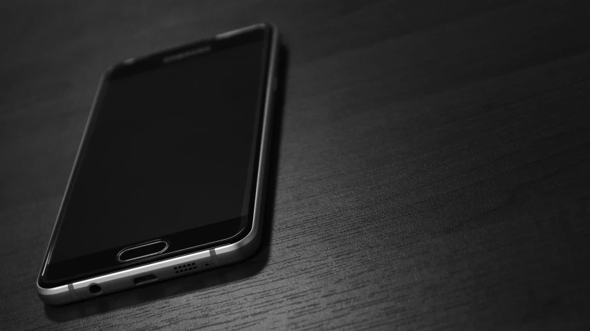 a cell phone sitting on top of a wooden table, a black and white photo, by Niko Henrichon, pixabay, realism, sharp sleek edged black armor, amoled wallpaper, android, gradient black to silver