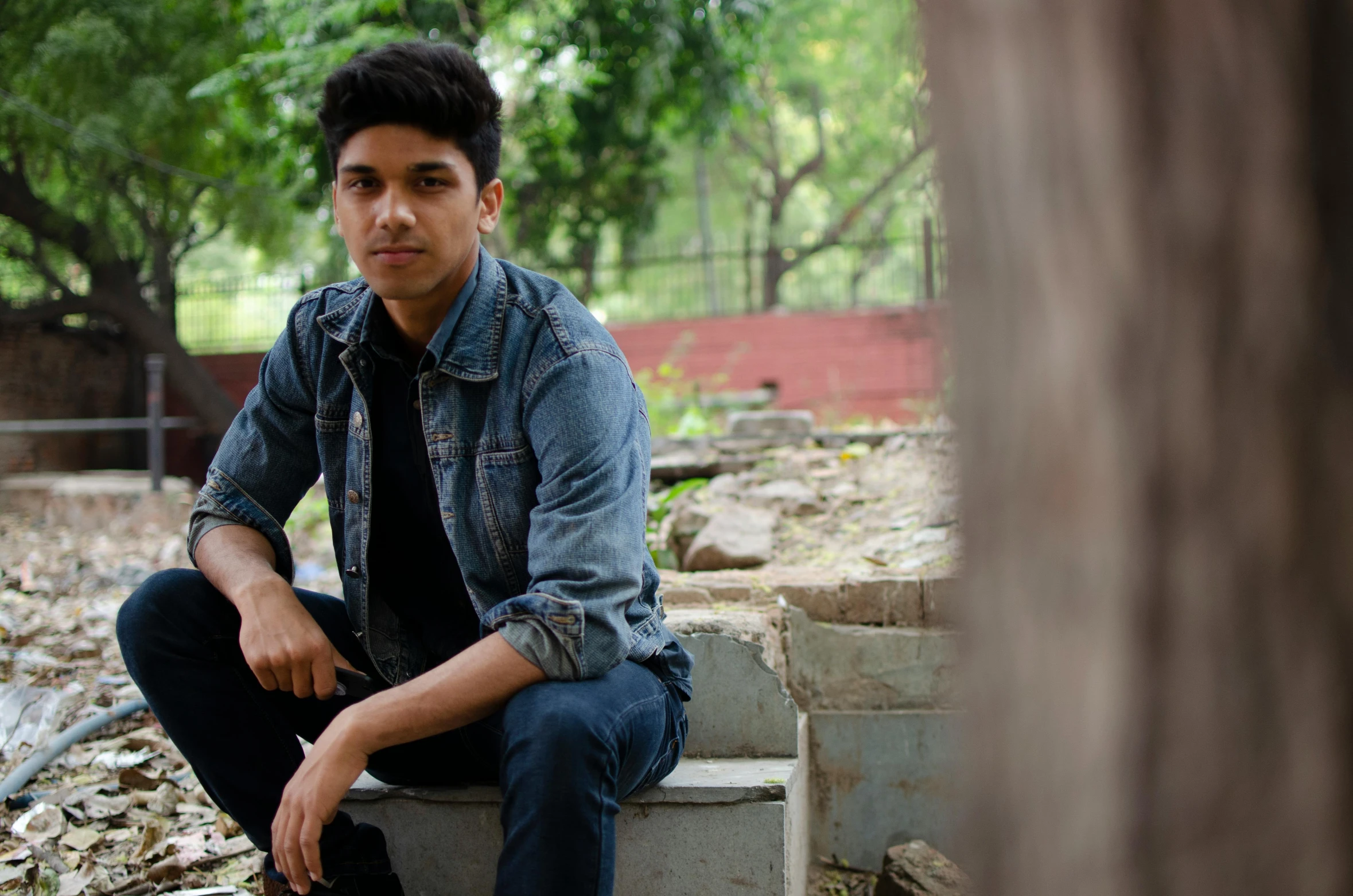 a young man sitting on top of a cement block, pexels contest winner, realism, a portrait of rahul kohli, trees in the background, wearing a jeans jackets, lgbtq