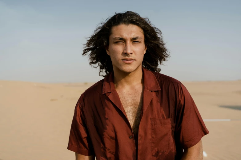 a man with long hair standing in the desert, trending on pexels, he looks like tye sheridan, middle eastern skin, red shirt, frank dillane