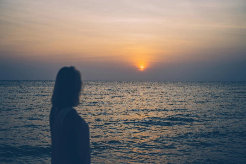 a woman standing on top of a beach next to the ocean, inspired by Elsa Bleda, pexels contest winner, happening, sunset halo behind her head, human staring blankly ahead, profile pic, slightly pixelated