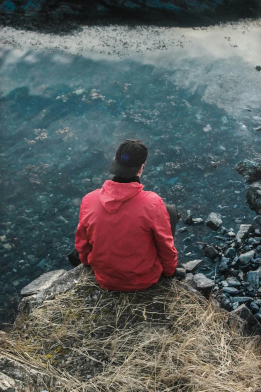 a man sitting on top of a rock next to a body of water, inspired by Elsa Bleda, pexels contest winner, wearing red jacket, sad look, backfacing, high angle shot