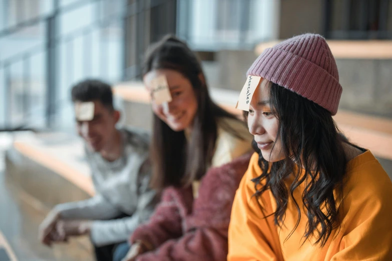 a group of people sitting next to each other on a bench, trending on pexels, mingei, wearing a pink head band, asian woman made from origami, black haired girl wearing hoodie, board games on a table