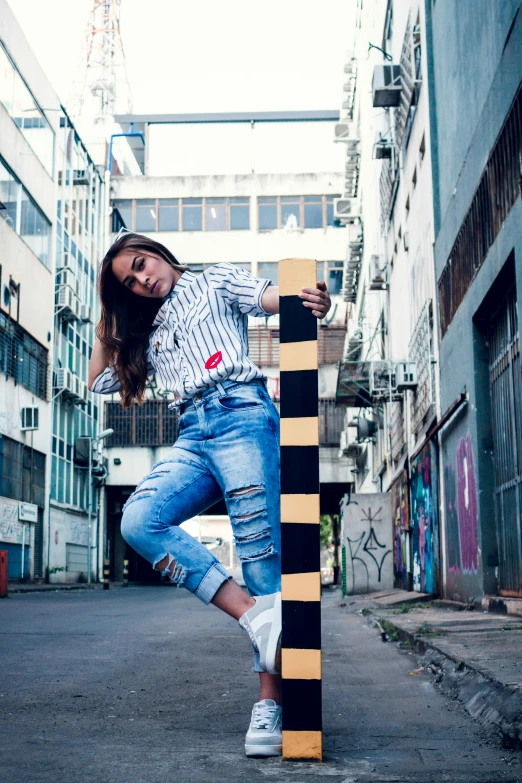 a woman leaning on a pole in the middle of a street, an album cover, inspired by Almeida Júnior, pexels contest winner, graffiti, confident action pose, denim, 15081959 21121991 01012000 4k, a wooden