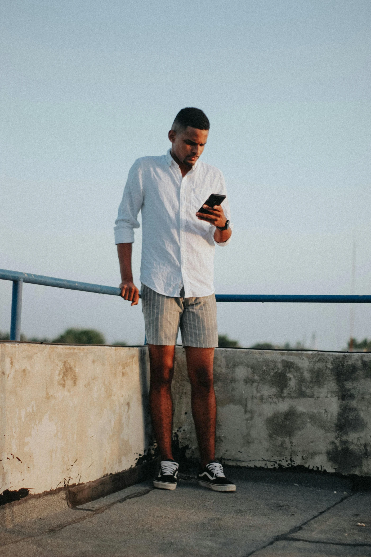 a man standing on a ledge looking at his cell phone, trending on unsplash, happening, dressed in a top and shorts, white and grey, brown skinned, tinder profile