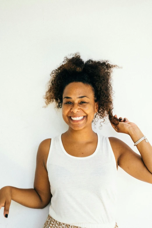 a woman standing in front of a white wall, natural hair, smiling playfully, pictured from the shoulders up, white backround