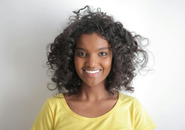 a young girl standing in front of a white wall, hurufiyya, dark short curly hair smiling, avatar image, close - up photograph, ( brown skin )