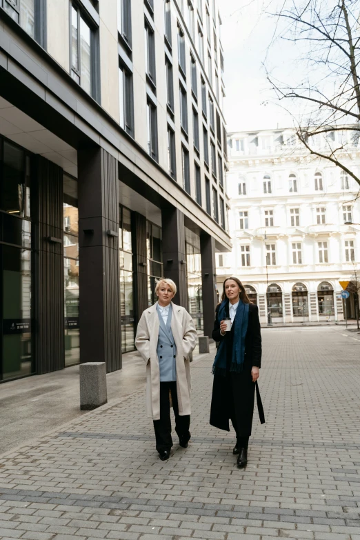 a couple of people walking down a sidewalk, wearing a long coat, vienna city, lesbian, full building