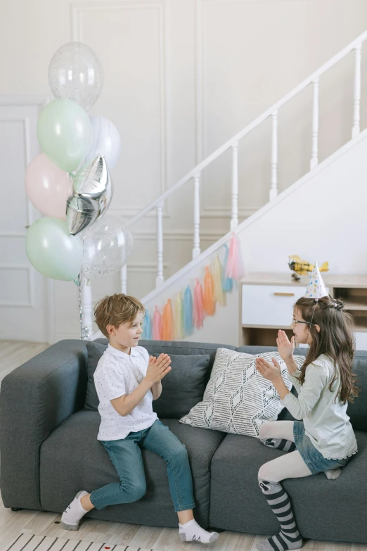 a couple of kids sitting on top of a couch, party balloons, silver and muted colors, promo image, waving