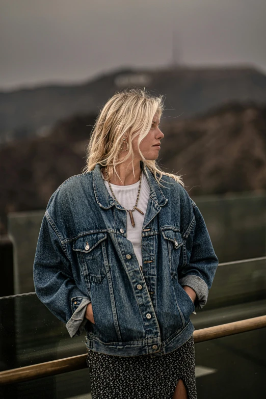 a woman standing on a balcony with mountains in the background, an album cover, trending on pexels, photorealism, denim jacket, blonde women, dressed in a ((ragged)), androgynous male