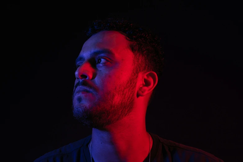 a man standing in front of a red light, with a black background, joel torres, red and blue black light, looking away from camera