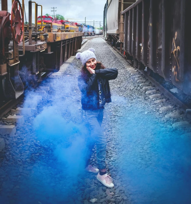 a woman standing next to a train on a train track, by Lucia Peka, pexels contest winner, graffiti, bright blue smoke, :6 smoke grenades, posing for a picture, cold color