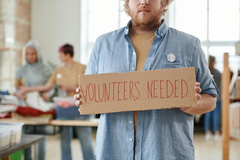 a man holding a sign that says volunteers needed, pexels contest winner, avatar image, colour photo, cardboard, looking from shoulder