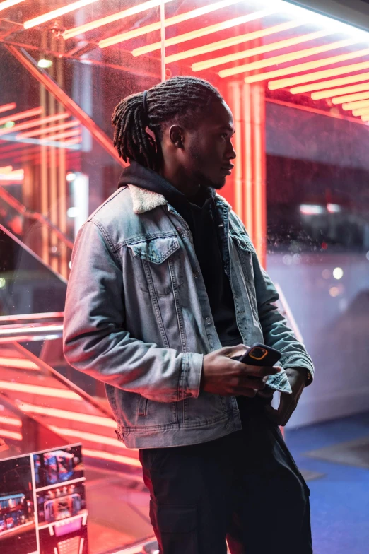 a man standing in front of a neon lit building, an album cover, trending on pexels, dreads, wearing a turtleneck and jacket, thoughtful ), looking at his phone