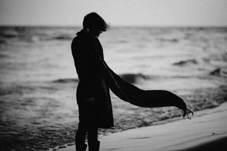 a person standing on a beach next to the ocean, a black and white photo, unsplash, romanticism, wrapped in a black scarf, declan mckenna, character silhouette, orelsan