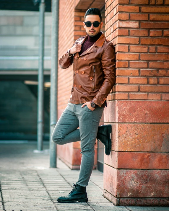 a man in a brown leather jacket leaning against a brick wall, by Cosmo Alexander, pexels contest winner, red brown and grey color scheme, full body picture, brown:-2, artem