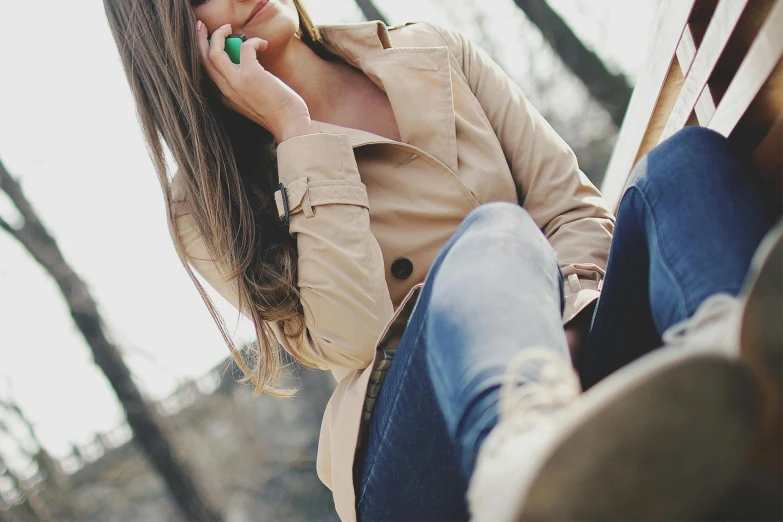 a woman sitting on a bench talking on a cell phone, trending on pexels, green and brown clothes, light brown trenchcoat, lipstick, ( ( ( wearing jeans ) ) )