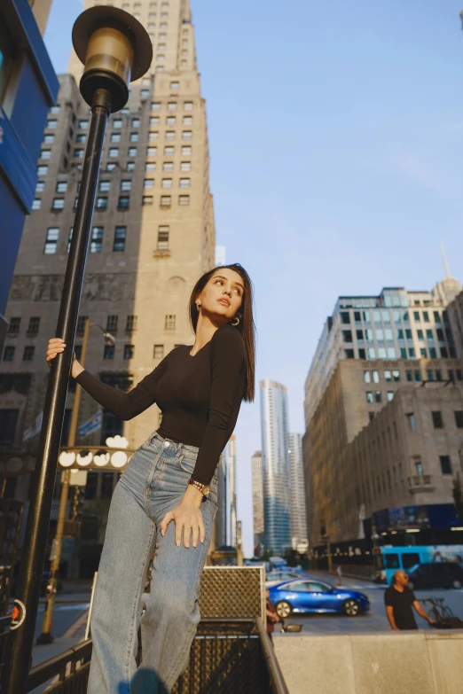 a woman standing next to a lamp post in a city, pexels contest winner, wearing jeans and a black hoodie, gal gadot china plate, modern chicago streets, tall buildings