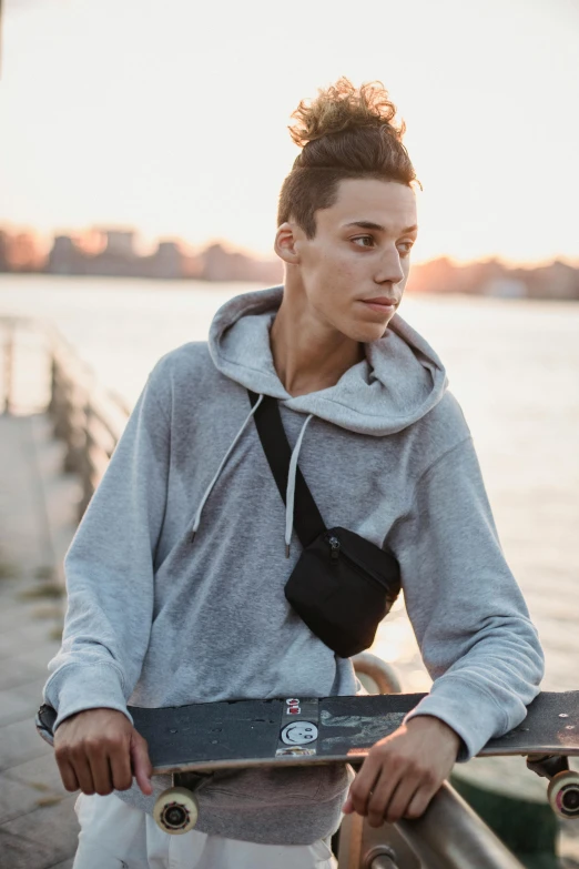 a young man holding a skateboard next to a body of water, grey hoodie, bag over the waist, jakub gazmercik, headshot
