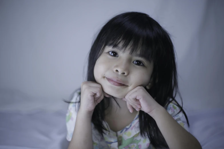 a little girl sitting on top of a bed, pexels contest winner, hurufiyya, detailed face of a asian girl, on a gray background, (night), small smile