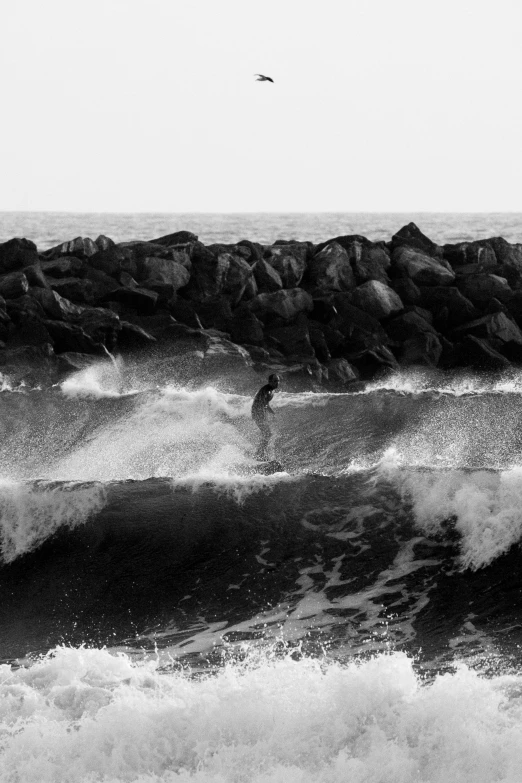 a man riding a wave on top of a surfboard, a black and white photo, by Dave Melvin, unsplash, conceptual art, with jagged rocks & eerie, joel meyerowitz, near a jetty, ((waves