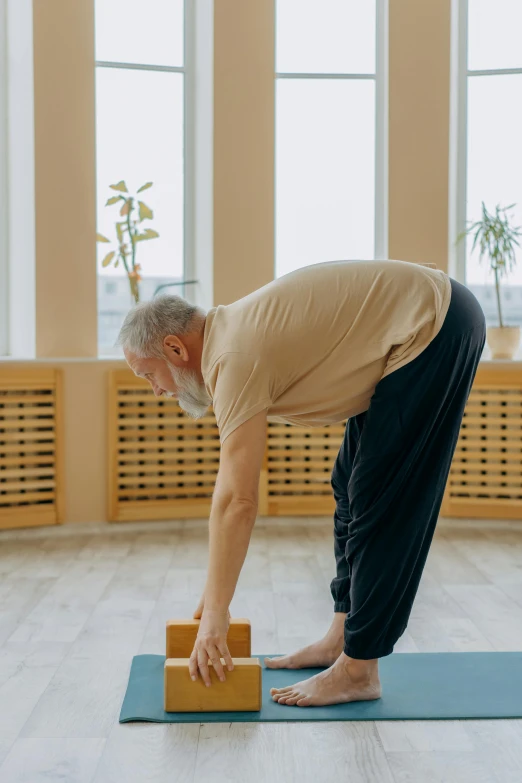 a man standing on a yoga mat with a block in his hand, nursing home, bending down slightly, square, profile image