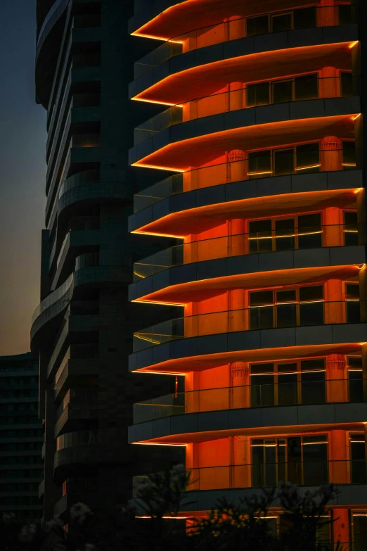 a tall building is lit up at night, inspired by Ricardo Bofill, flickr, orange ribbons, curvy build, afternoon light, crenellated balconies