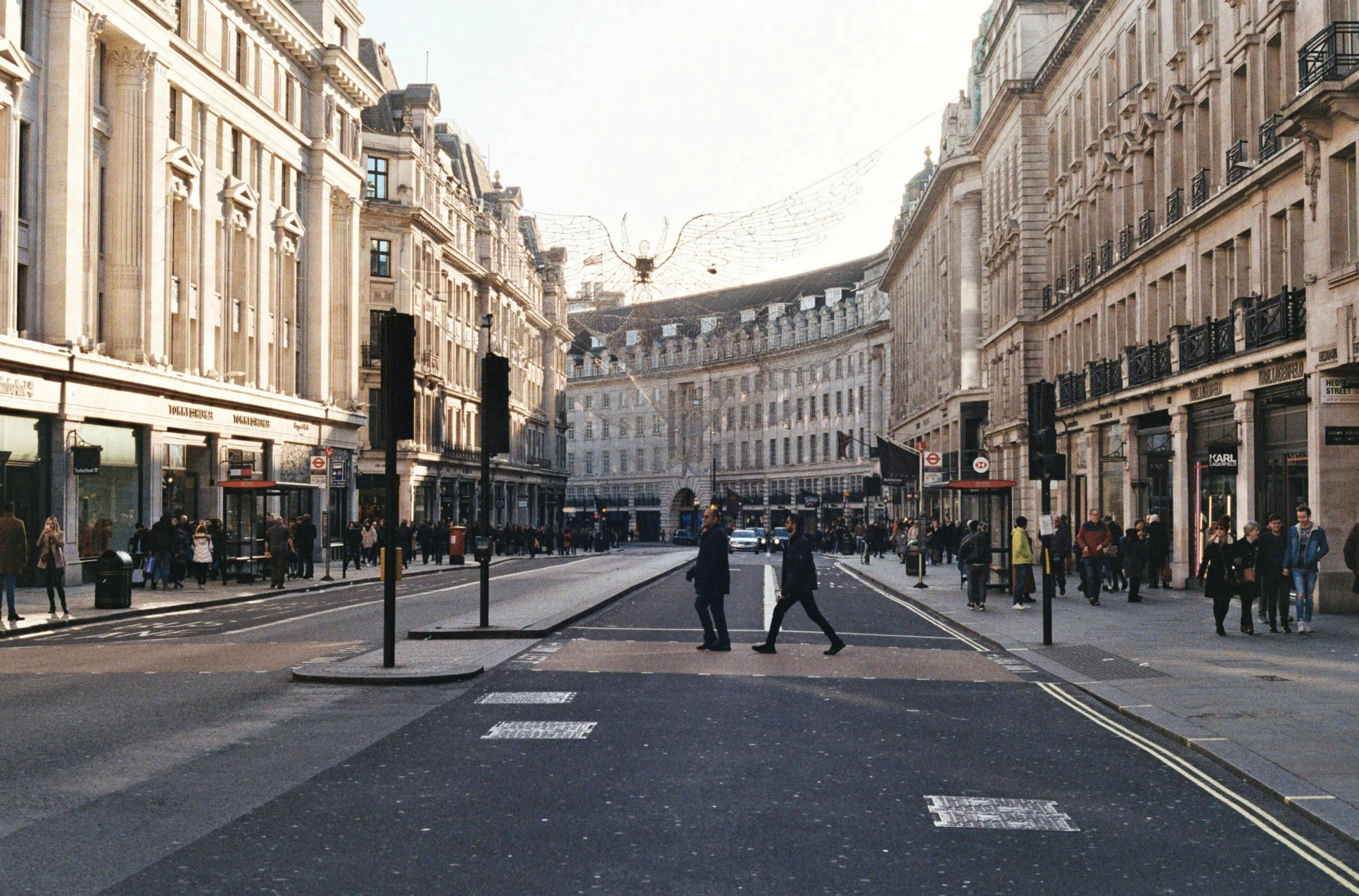 a group of people walking down a street next to tall buildings, an album cover, by Nina Hamnett, pexels contest winner, renaissance, on a great neoclassical square, united kingdom, intersection, full daylight