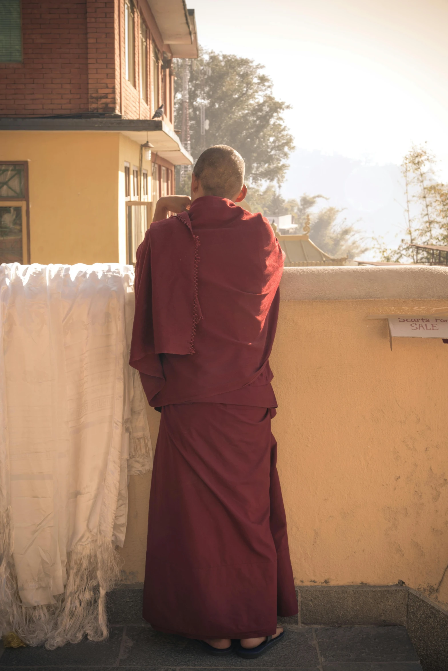 a man in a red robe leaning against a wall, inspired by Steve McCurry, trending on unsplash, bhutan, back view, sunny day, wearing jedi robes and a sari