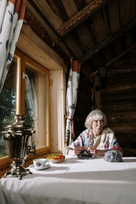 a woman sitting at a table in front of a window, by Eero Järnefelt, unsplash, fine art, log cabin, traditional russia, in an igloo, older woman