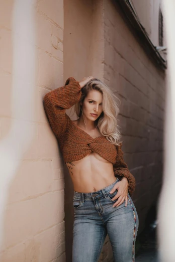a woman leaning against a wall with her hands on her hips, by Robbie Trevino, trending on pexels, realism, beautiful blonde girl, brown sweater, croptop, fit pic