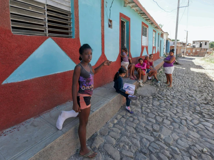 a group of people sitting on the side of a building, by Ceferí Olivé, happening, village girl reading a book, street of teal stone, walking towards the camera, 2018