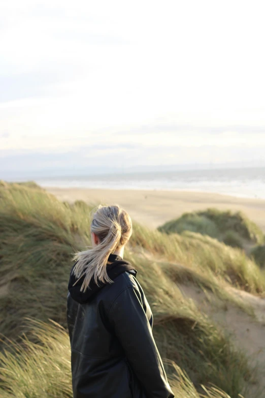 a woman standing on top of a sandy beach, by Maria van Oosterwijk, unsplash, long grass in the foreground, looking off into the distance, coastline, university