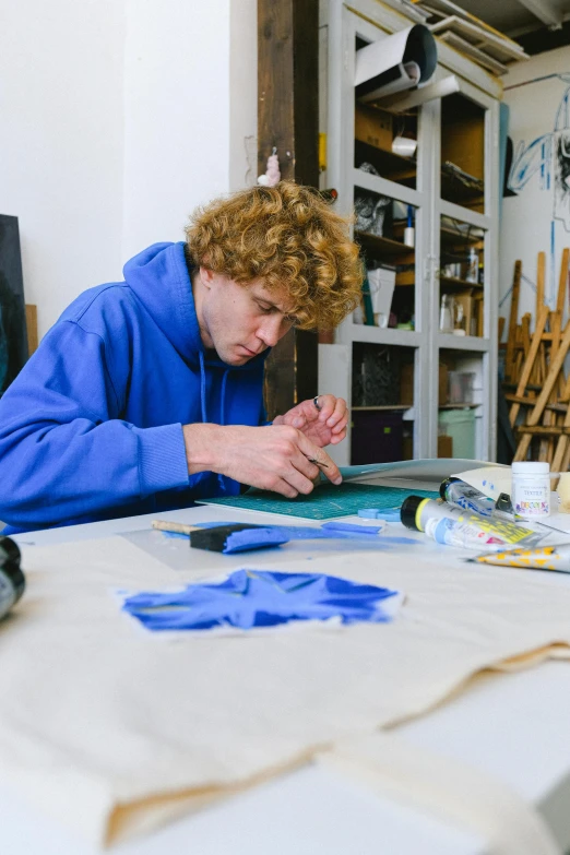 a man sitting at a table working on a piece of art, wearing a blue hoodie, teenager, caspar david, paint knife