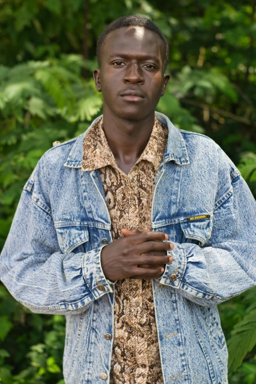 a man standing in front of a lush green forest, an album cover, pexels, renaissance, adut akech, denim jacket, patterned clothing, portait image