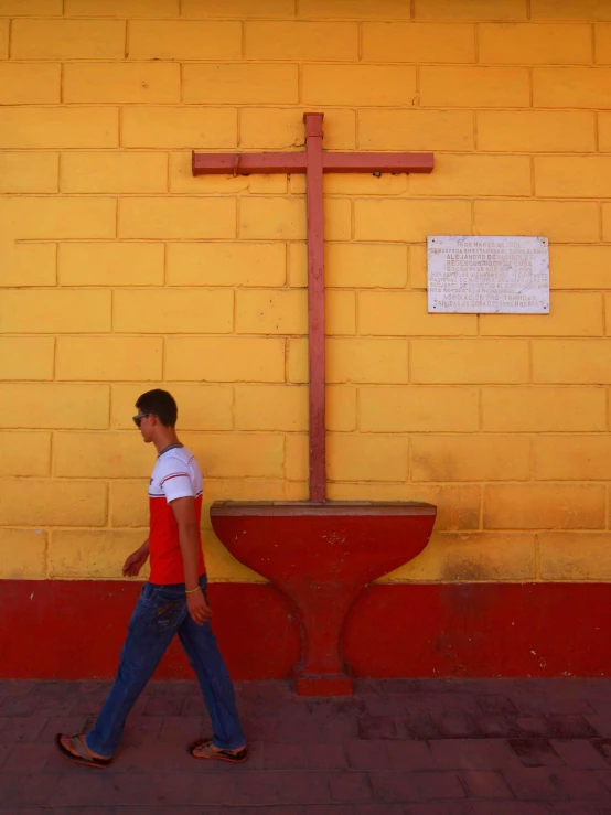 a man walking past a wall with a cross on it, yellow and red, karla ortiz, in town, ap news photograph