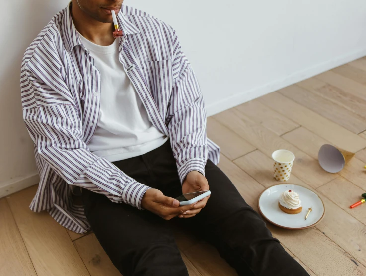 a man sitting on the floor looking at his cell phone, by Nina Hamnett, trending on pexels, happening, striped shirt, having a snack, button - up shirt, kousuke oono