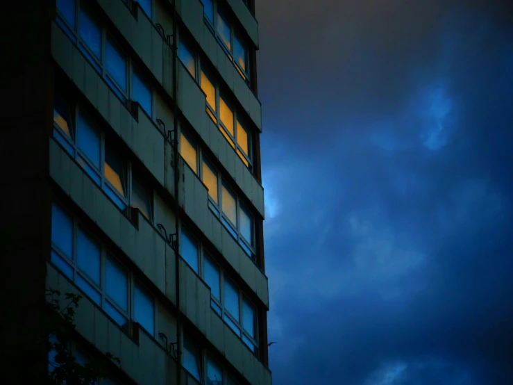 a tall building sitting under a cloudy sky, an album cover, inspired by Elsa Bleda, unsplash, brutalism, window to night time, yellow and blue, vibrant but dreary blue, late summer evening