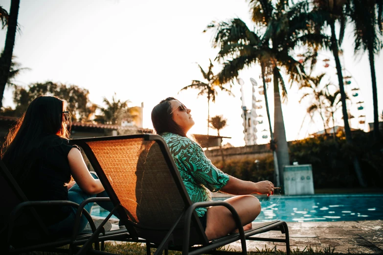 two women sitting in lawn chairs next to a swimming pool, pexels contest winner, happening, sun behind her, avatar image, high quality image, palm trees in the background