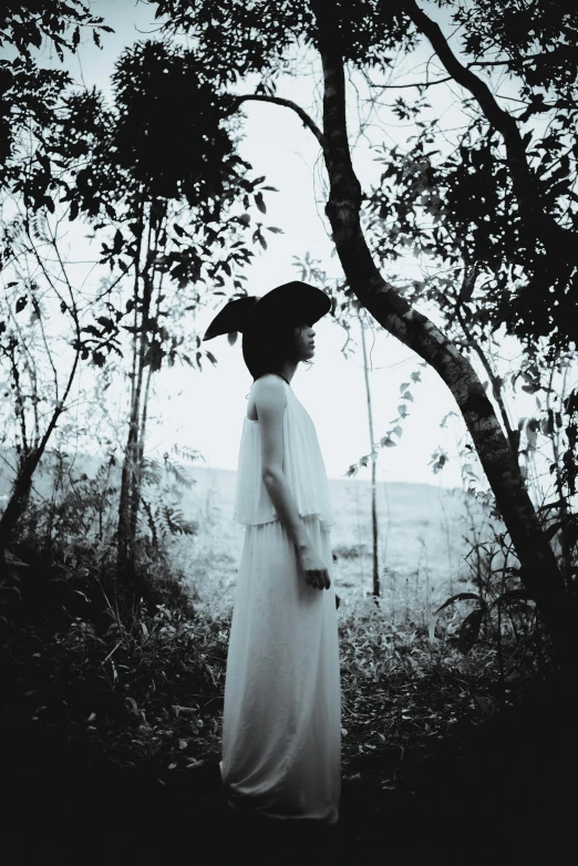 a woman in a white dress standing in the woods, a black and white photo, unsplash, magical realism, vietnamese woman, wearing a witch hat, taken in the late 2010s, medium format. soft light