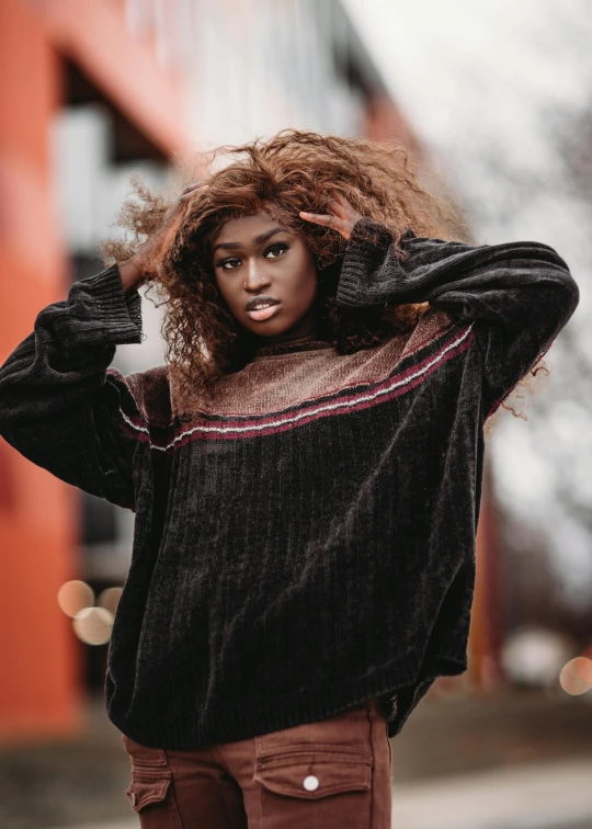 a woman with curly hair standing in front of a building, trending on pexels, brown sweater, ( ( dark skin ) ), non binary model, maiden with copper hair