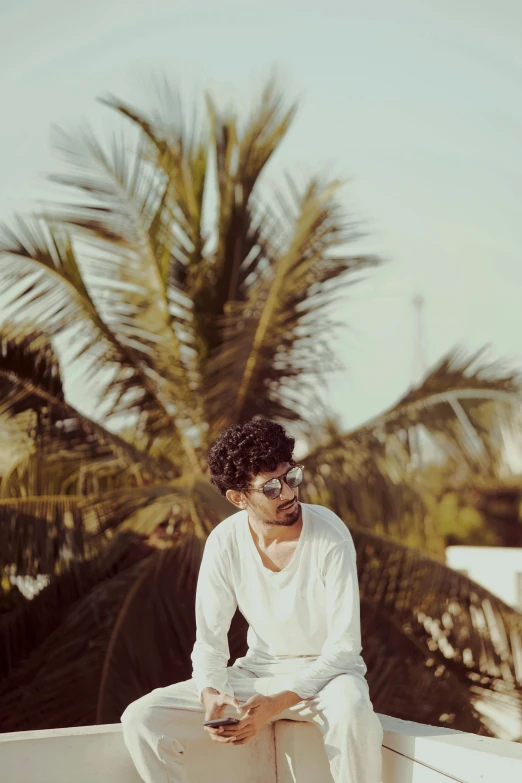 a man sitting on a ledge next to a palm tree, an album cover, by Max Dauthendey, pexels contest winner, curly haired, vinayak, profile image, with sunglass