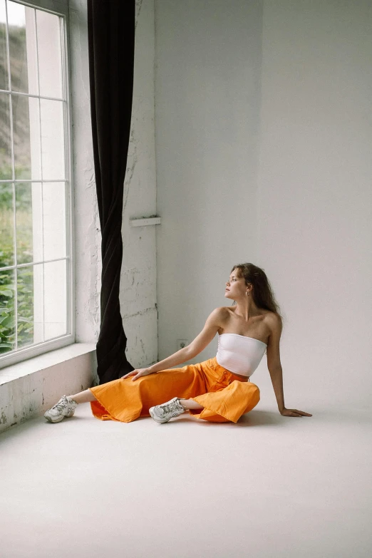 a woman sitting on the floor in front of a window, inspired by Elsa Bleda, trending on pexels, happening, white and orange, portrait sophie mudd, pants, in front of white back drop