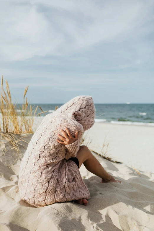 a woman sitting on top of a sandy beach, by Matija Jama, trending on pexels, wearing a pastel pink hoodie, cozy under a blanket, devastated, wearing a cardigan