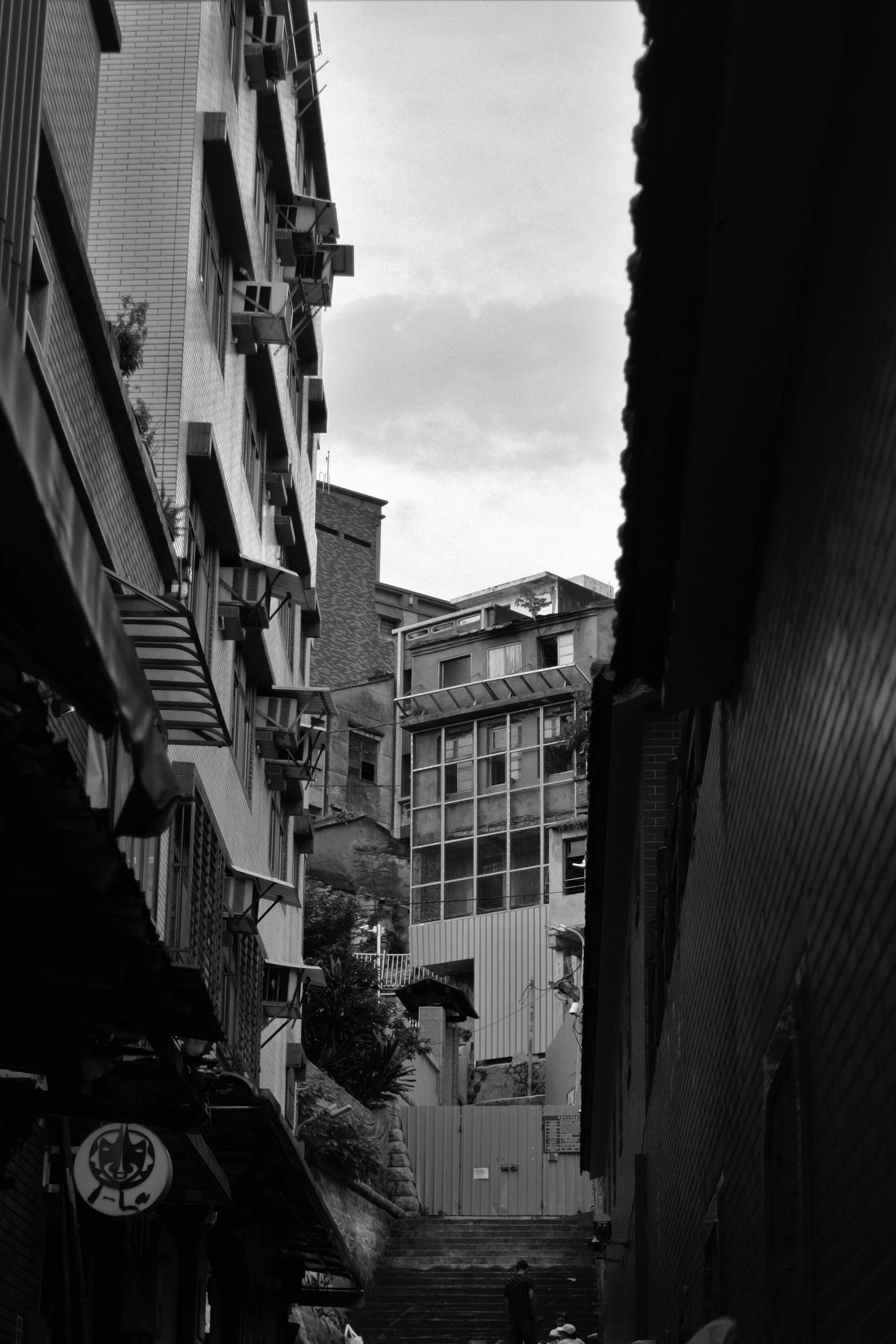 a man riding a skateboard down a street next to tall buildings, a black and white photo, inspired by Thomas Struth, unsplash, mingei, in a narrow chinese alley, photo taken from a boat, location ( favela ), looking out