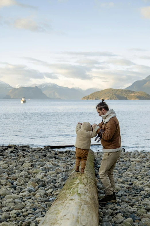 a woman standing on top of a log next to a body of water, with a kid, mountains and oceans, rocky beach, father with child