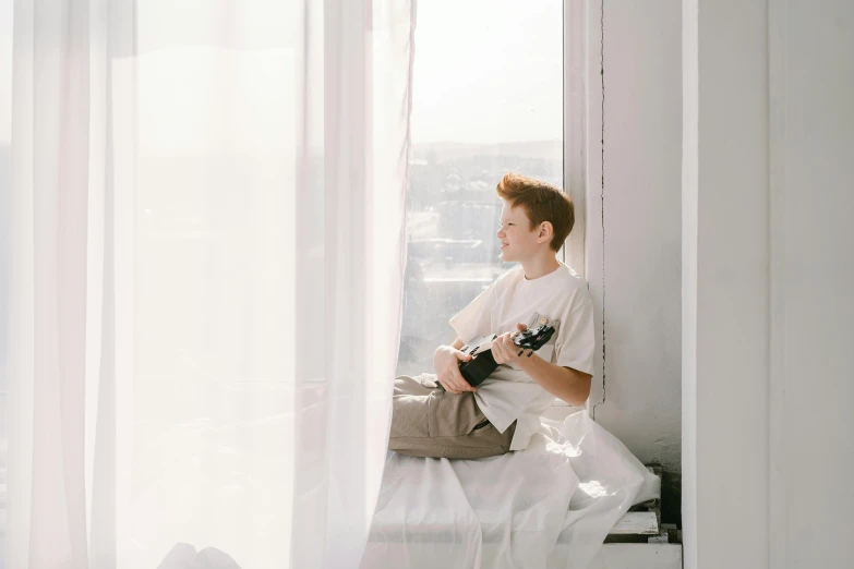 a man sitting on top of a bed next to a window, by Emma Andijewska, pexels contest winner, ukulele, red haired teen boy, dressed in a white t shirt, 1 4 9 3