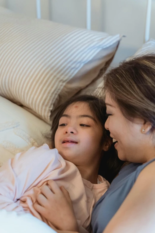 a woman laying on top of a bed next to a little girl, smiling at each other, asian descent, comforting, lgbtq