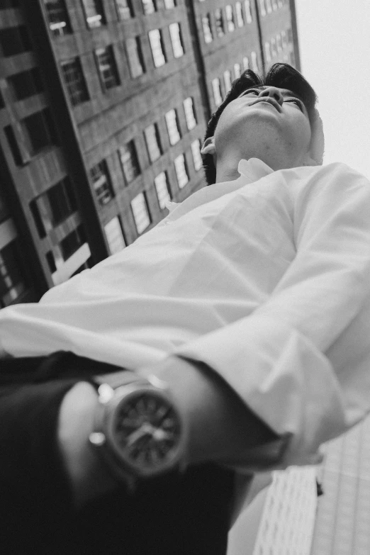 a man standing in front of a tall building, a black and white photo, unsplash, realism, wearing a watch, lying down, clothed in white shirt, sittin